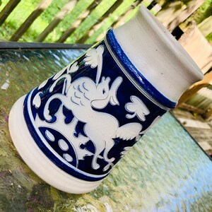 May include: A white ceramic stein with a blue and white design of a mythical creature with wings. The stein has a blue rim and a white base.