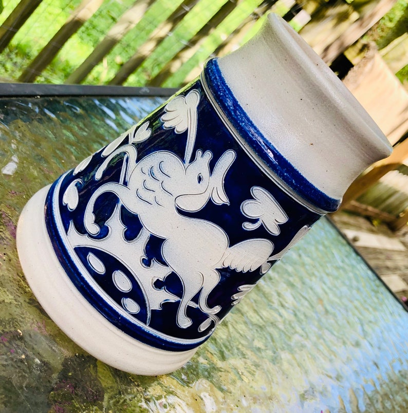 May include: A white ceramic stein with a blue and white design of a mythical creature with wings. The stein has a blue rim and a white base.