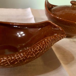 May include: A close-up of two brown ceramic serving dishes. One is a gravy boat with a textured rim, and the other is a covered dish. Both have a glossy finish. The dishes are on a white surface.