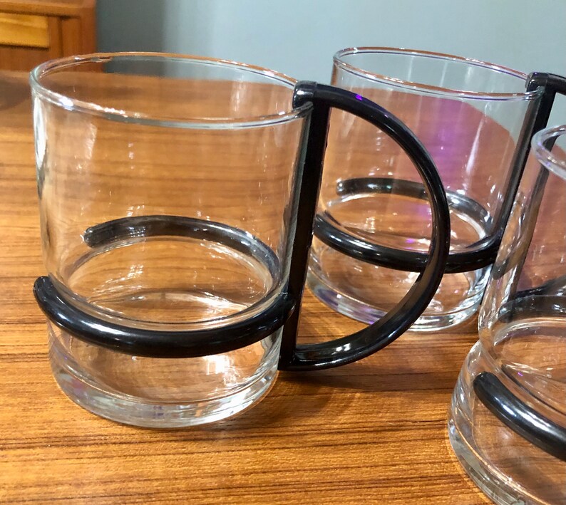 Op de afbeelding: Vier glazen glazen met zwarte kunststof handvatten. De glazen zijn met elkaar verbonden door de handvatten.