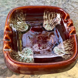 May include: A square ceramic ashtray with a brown glaze and white and green speckled accents. The ashtray has four indentations for cigarettes.