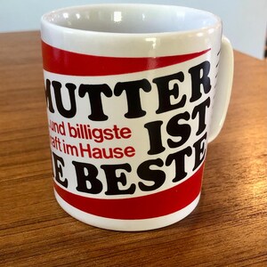 May include: White ceramic mug with a red and black design. The text on the mug reads "Mutter ist die Beste" and "und billigste Kraft im Hause".