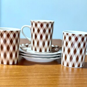 May include: Three white ceramic mugs with a brown and white diamond pattern, along with matching saucers and spoons. The mugs have a curved handle and are set on a wooden surface.