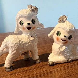 May include: Two ceramic lamb figurines. One lamb is standing, the other is sitting. Both are white with textured fur, green eyes, and rosy cheeks. They have small, decorative floral accents on their heads and chests.