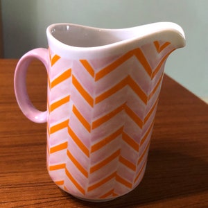 May include: A white ceramic pitcher with a pink handle and a chevron pattern in orange and pink.