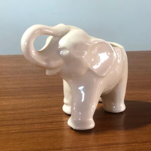 May include: A ceramic elephant planter in a light pink color. The elephant has its trunk curled up and its ears are large and floppy.