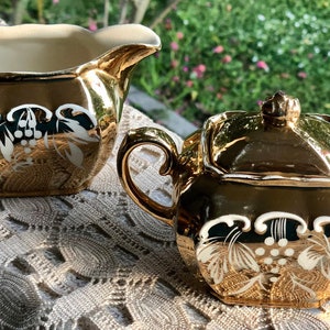 May include: A gold-colored creamer and covered sugar bowl set. The set features a white grape and leaf design. The creamer has a curved spout and handle. The sugar bowl has a lid with a decorative finial and a handle.