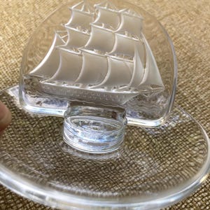 May include: Clear glass decorative dish with a detailed sailboat design. The sailboat is etched into the glass, featuring multiple sails and a detailed hull. The dish has a circular base and a raised, curved top.