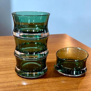 May include: A set of three green glass tumblers and a small bowl. The tumblers are stacked, with a unique bamboo-like design. The bowl has a curved shape. The glass has a reflective quality, and the set is on a wooden surface.