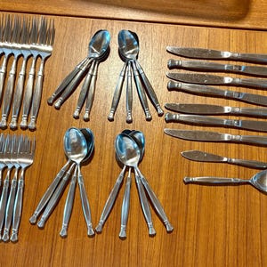 May include: A collection of stainless steel silverware, including forks, spoons, and knives, arranged on a wooden surface. The silverware has a simple, elegant design, perfect for a formal dining setting.