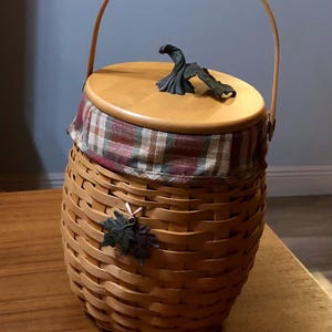 May include: A woven wicker basket with a wooden lid and a plaid fabric lining. The lid has a decorative metal pumpkin stem on top. The basket is brown and has a handle.