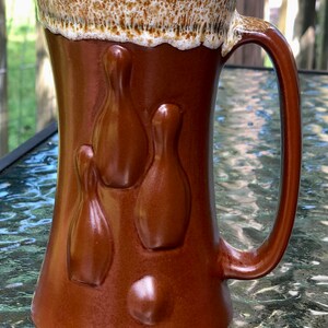 May include: A brown ceramic mug with a white rim and a design of bowling pins. The mug has a handle.