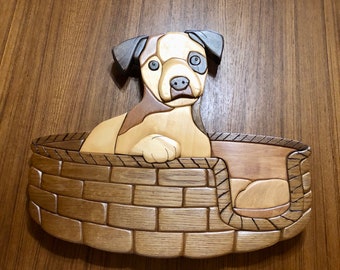 Op de afbeelding: Een houten sculptuur van een hond in een hondenbed. De hond is bruin en wit met zwarte ogen en een roze neus. Het hondenbed is gemaakt van hout en heeft een baksteenpatroon.