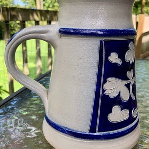 May include: A white ceramic beer stein with a blue and white floral design. The stein has a handle and a wide base.