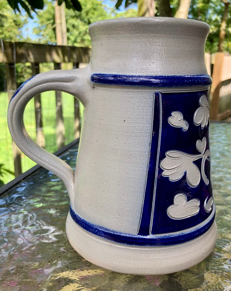 May include: A white ceramic beer stein with a blue and white floral design. The stein has a handle and a wide base.