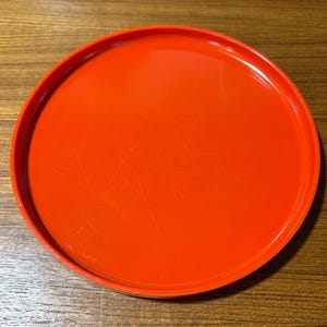 May include: A round, orange plastic tray with a slightly raised edge. The tray has visible scratches on its surface, indicating prior use. The bright orange color provides a pop of color.