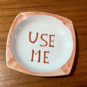 May include: A square ceramic ashtray with a peach-colored rim and the words "USE ME" written in brown on the white surface.