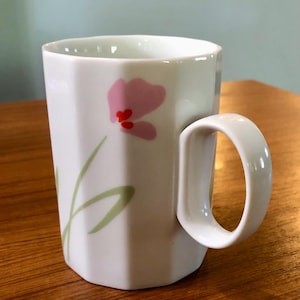 May include: White ceramic mug with a pink flower and green leaves design. The mug has a hexagonal shape and a white handle.