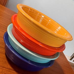 May include: A stack of colorful ceramic oval baking dishes in shades of yellow, orange, turquoise, and blue. The dishes are stacked at an angle, showcasing their vibrant colors and smooth, glossy finish. The dishes are on a wooden surface.
