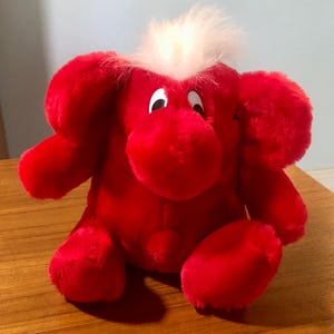 May include: A plush red stuffed animal with a white tuft of hair on top. The toy has large, round ears, a rounded snout, and small black and white eyes. It is sitting on a wooden surface.