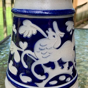 May include: A white ceramic beer stein with a blue glaze and a white design of a bird with wings and a floral pattern.