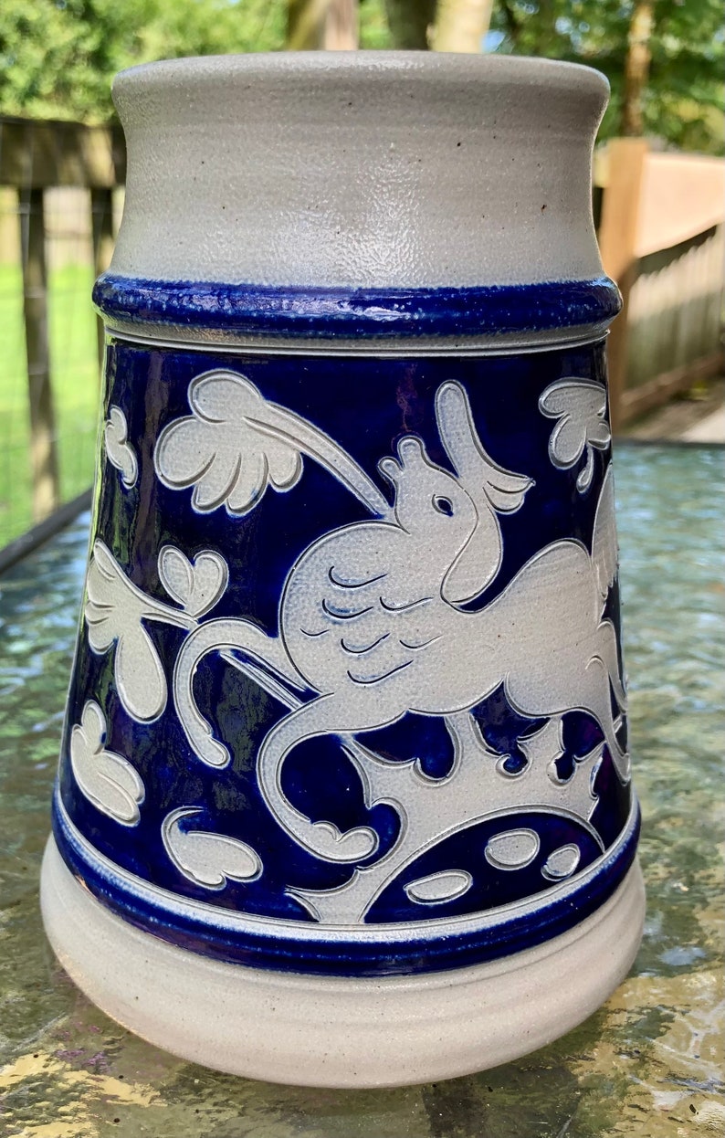 May include: A white ceramic beer stein with a blue glaze and a white design of a bird with wings and a floral pattern.