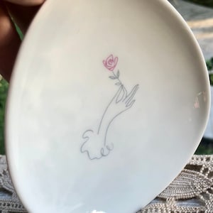 May include: A white ceramic dish with a stylized drawing of a hand holding a single pink rose.