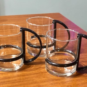 May include: Three clear glass mugs with black plastic handles. The mugs have a wide, cylindrical shape and are perfect for enjoying a hot beverage.