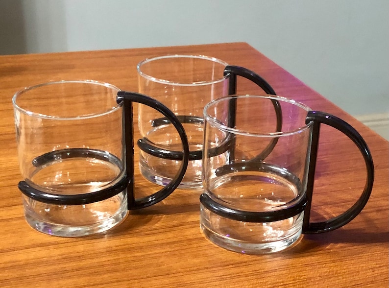 Op de afbeelding: Drie heldere glazen bekers met zwarte kunststof handvatten. De bekers hebben een brede, cilindrische vorm en zijn perfect om van een warme drank te genieten.