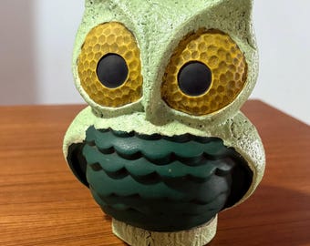 Op de afbeelding: Een keramieken uilfiguur met een getextureerd, lichtgroen lichaam en grote, gele ogen met zwarte pupillen. De borst van de uil is donkergroen met een geschulpte vormgeving. Het is een decoratief item.