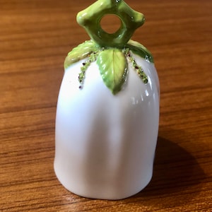 May include: White ceramic bell with a green leaf design and a small hole in the top. The bell has a rounded shape and a smooth surface.