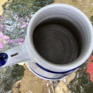 May include: A white ceramic beer stein with a blue and white design. The stein has a handle and a wide mouth.