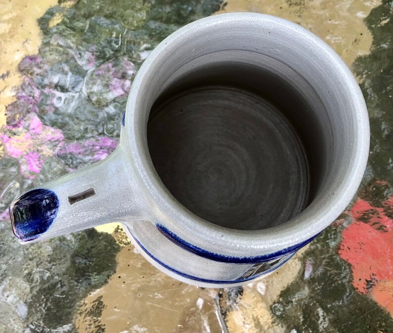 May include: A white ceramic beer stein with a blue and white design. The stein has a handle and a wide mouth.