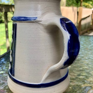 May include: A white ceramic beer stein with a blue handle and blue trim. The stein features a traditional German design with white dots and a blue and white pattern.