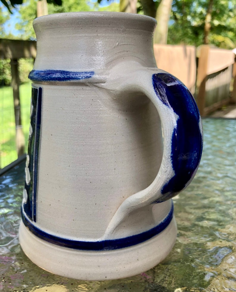 May include: A white ceramic beer stein with a blue handle and blue trim. The stein features a traditional German design with white dots and a blue and white pattern.
