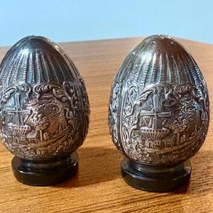 May include: Two antique silver-colored salt and pepper shakers. The egg-shaped shakers have a detailed embossed design featuring a landscape with buildings and foliage. They sit on black bases and are displayed on a wooden surface.