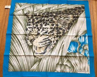 May include: A blue silk scarf with a white background featuring a detailed illustration of a leopard in a jungle setting. The scarf has a blue border and the text "Salvatore Ferragamo" is printed in the bottom right corner.