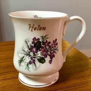 May include: White ceramic mug with a gold rim and a floral design featuring purple flowers and a white ribbon. The mug has the name "Helen" written in gold lettering.