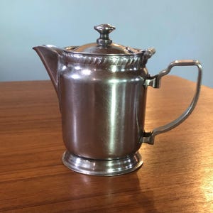 May include: A silver stainless steel creamer or gravy pitcher with a lid and a handle. The pitcher has a decorative band around the top.