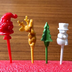 May include: Four festive plastic food picks, each with a different Christmas theme: a red Santa Claus, a yellow reindeer, a green Christmas tree, and a white snowman.