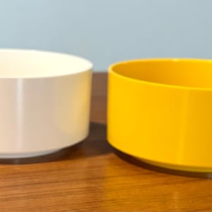 May include: Two cylindrical containers. One is cream-colored, the other is bright yellow. Both have a low profile and a slightly flared base. They sit on a wooden surface, with a light blue background.