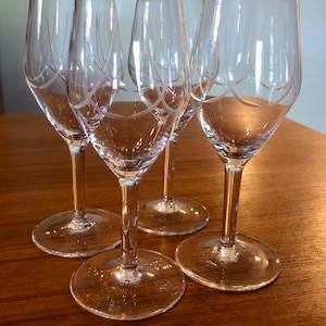 May include: Four clear wine glasses with etched designs. The glasses have a tall, slender stem and a wide base. The etched design is a series of curved lines near the top of the glass. The glasses are arranged on a wooden surface.