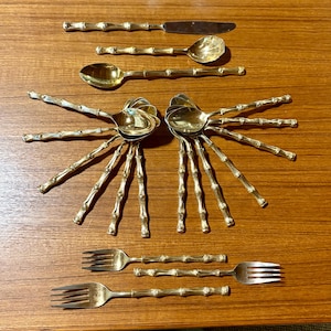 May include: A collection of gold-colored bamboo-style silverware, including spoons, forks, and a knife. The silverware is arranged on a wooden surface. The spoons have a unique design, with multiple bamboo-style stems radiating from the bowl.