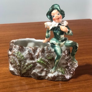 May include: A ceramic planter shaped like a rock with a green and white elf sitting on top. The elf is wearing a green hat and has a white collar. The planter has a brown and green glaze.
