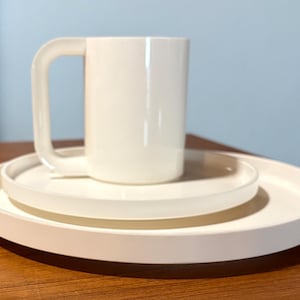 May include: A white mug with a square handle rests on a stack of three white plates. The plates are different sizes, with a large, round plate at the bottom and a smaller plate on top. The mug and plates are made of a solid, opaque material.