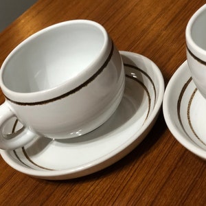 May include: Two white ceramic coffee cups with brown stripes around the rim. The cups are sitting on matching saucers with brown stripes.