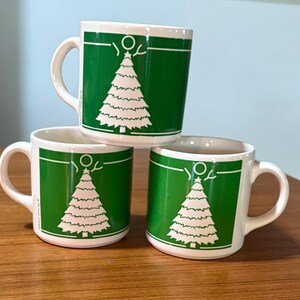 May include: Three white ceramic mugs stacked on a wooden surface. Each mug features a green panel with a white Christmas tree design. The mugs have white handles and a white rim.