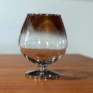 May include: A clear brandy snifter with a silver-colored base and rim. The glass has a wide bowl and a short stem. The glass is sitting on a wooden surface.