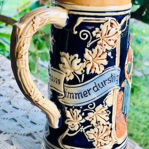 May include: A blue and white ceramic beer stein with a handle shaped like a vine. The stein has a brown rim and base and features a scene of a woman holding a pitcher and a banner that reads "immer durstig" with the year 2029.