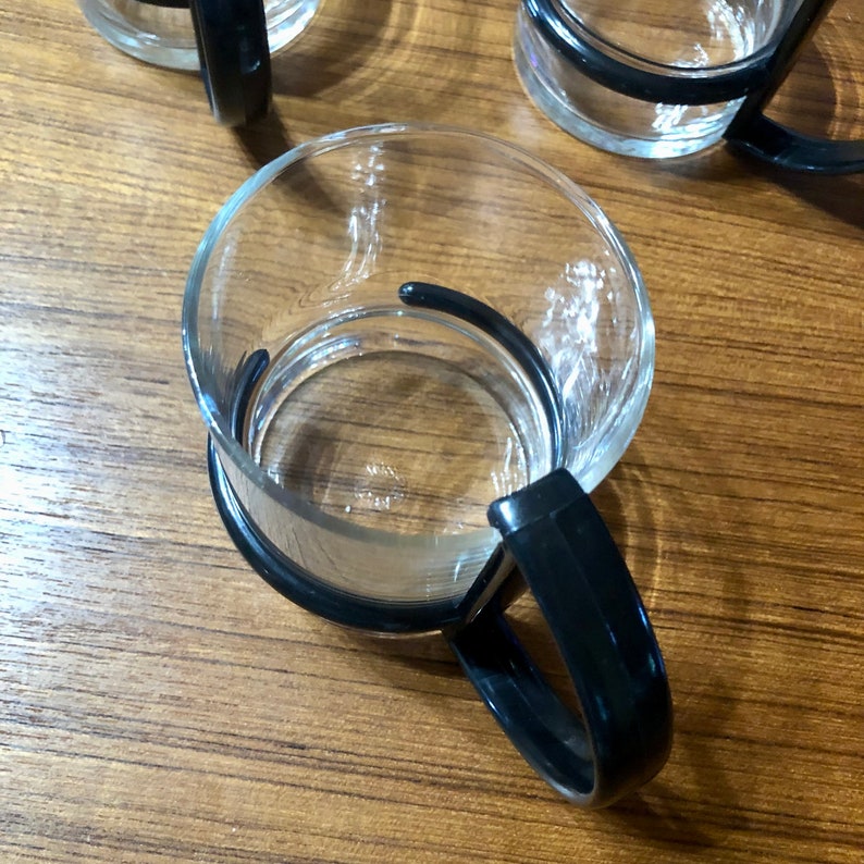 Op de afbeelding: Een heldere glazen beker met een zwarte kunststof handvat. De beker is leeg en staat op een houten oppervlak.